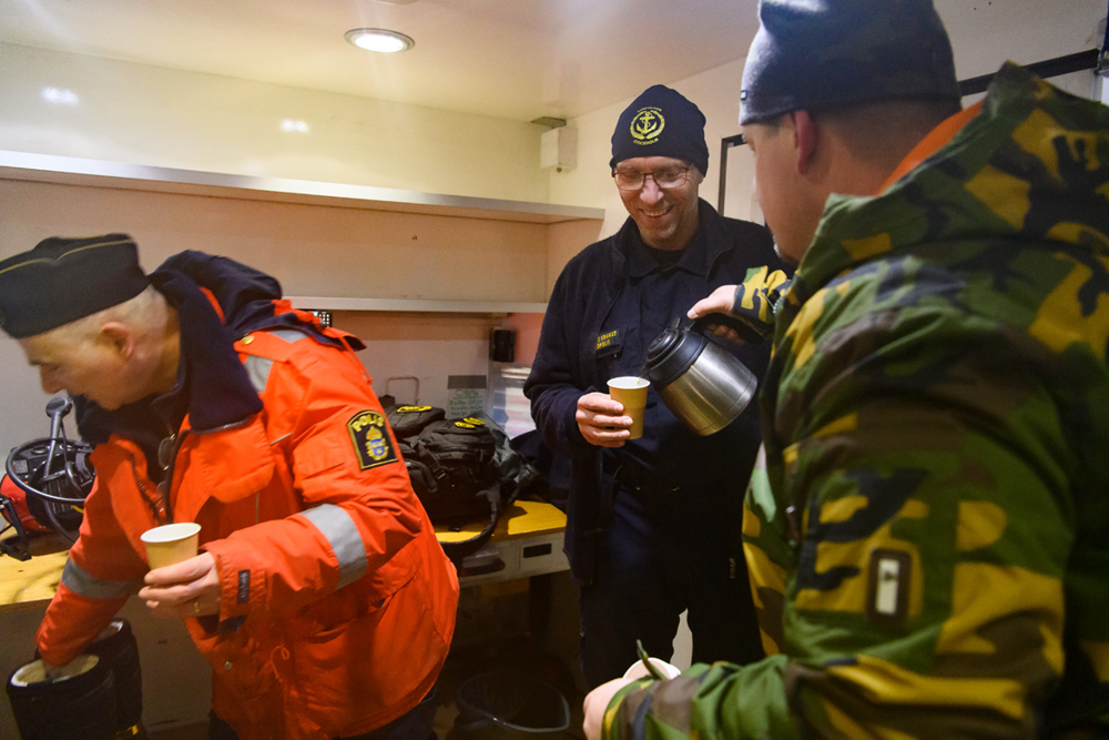 En dykare håller i sin kollegas luftslang ovanför is medan kollegan dyker.  I förgrunden ses dykledaren. Foto.