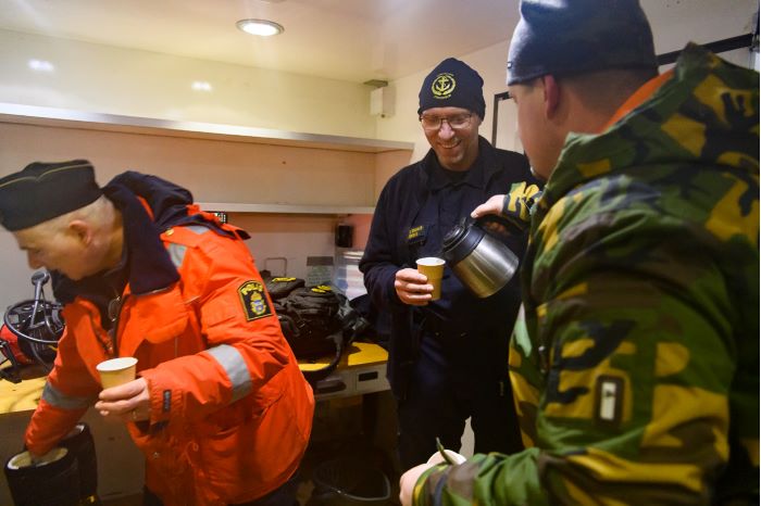 En dykare håller i sin kollegas luftslang ovanför is medan kollegan dyker.  I förgrunden ses dykledaren. Foto.