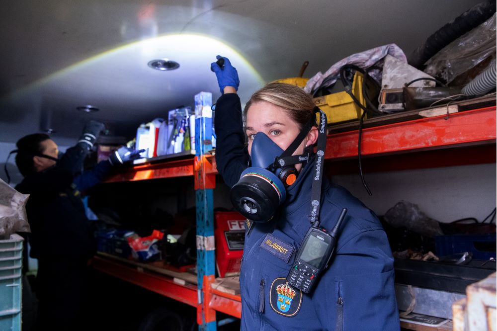 Miljöutredaren Ellen Forsberg står med skyddsmask i en container och lyser med en ficklampa bort mot sin utredarkollega Erik Wikstrand.