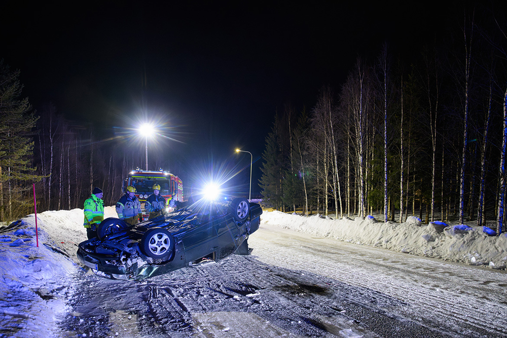 En voltad bil ligger med taket ned på den snöiga vägen och räddningstjänsten står bredvid. Foto..