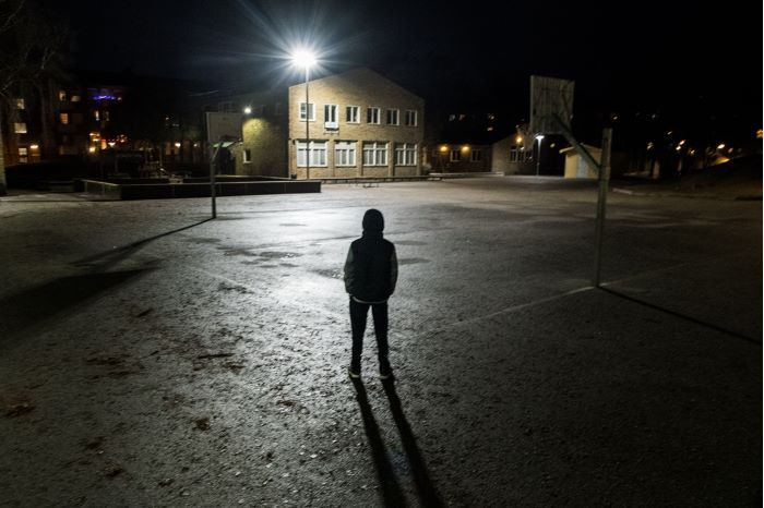 En ensam ung kille i huvtröja står i mörkret på en skolgård. Foto.