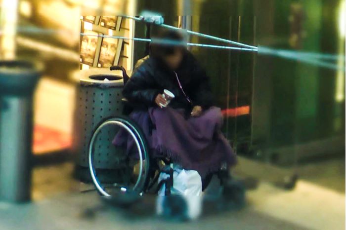 En person sitter i rullstol utanför en butik, med en pappersmugg i handen. Bilden är suddad för att skydda identiteten på personen i bilden.