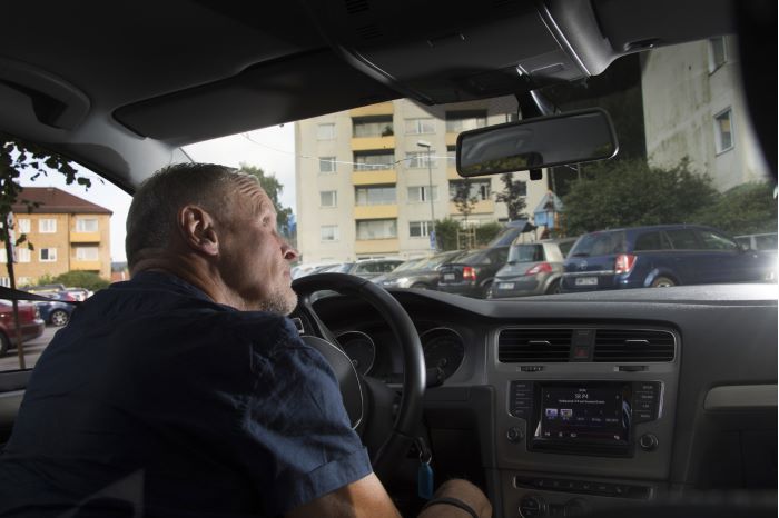 Mats Paulsson sitter i förarsätet på en bil och spanar ut genom vindrutan mot ett höghusområde.
