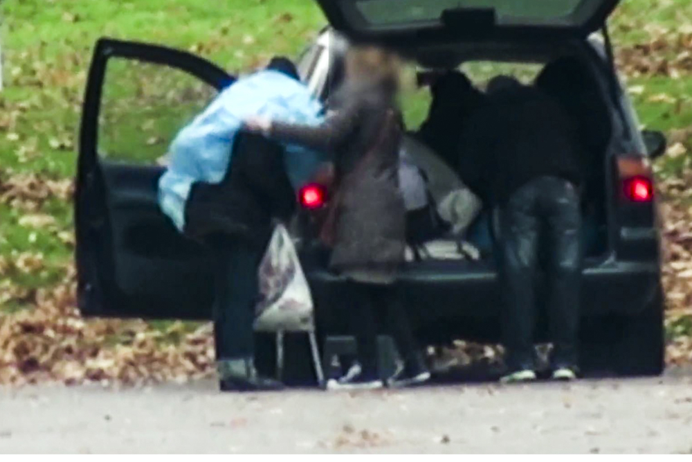 Tre personer står bakom en bil, som har bagageluckan öppen. Ena personen får en blå plastponcho över axlarna. Bilden är suddad för att skydda personernas identitet.