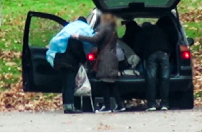 Tre personer står bakom en bil, som har bagageluckan öppen. Ena personen får en blå plastponcho över axlarna. Bilden är suddad för att skydda personernas identitet.