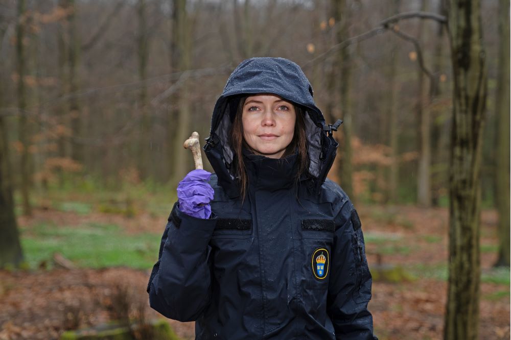 Clara Alfsdotter står i skogen med jackhuvan över huvudet och håller i en benknota med en plasthandske på handen. Foto.