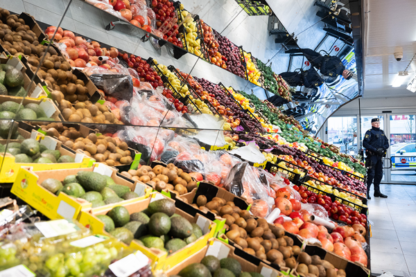 En polis ses stå vid ingången till butiken och i förgrunden ligger hyllor med olika sorters frukt. Foto.