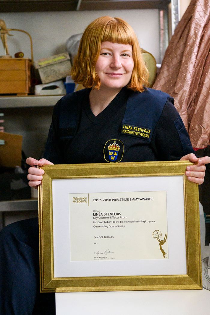 Linéa Stenfors sitter och håller i ett inramat diplom för en Emmy Award för sitt kostymarbete. Foto.