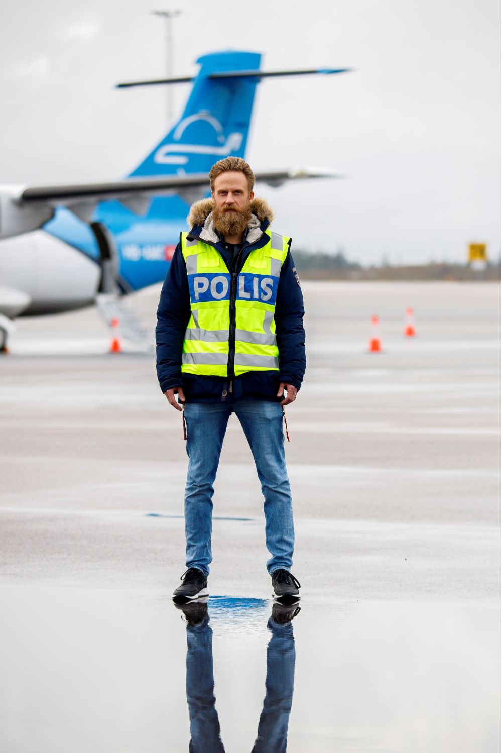 Verkställaren Andreas står iklädd gul  reflexväst som det står "polis" på ute på en landningsbana med ett flygplan i bakgrunden. Foto.