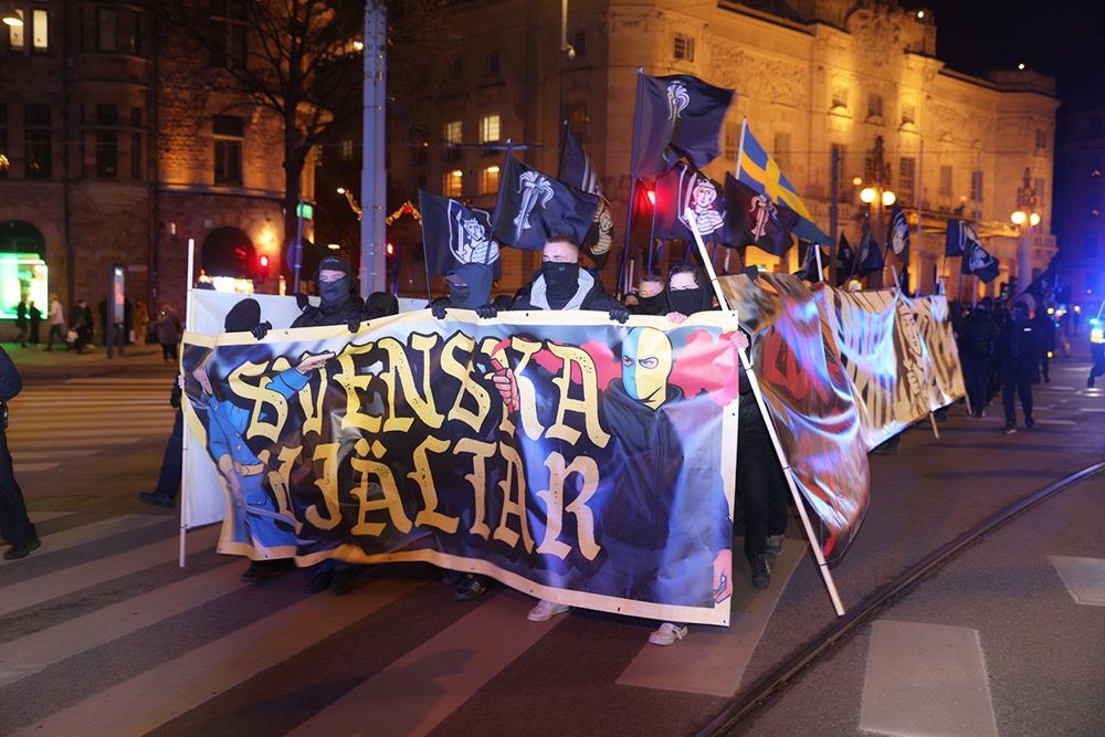 Maskerade personer går i en manifestation med symboler från en aktivklubb och i fronten en banderoll där det står "Svenska hjältar". Foto. 