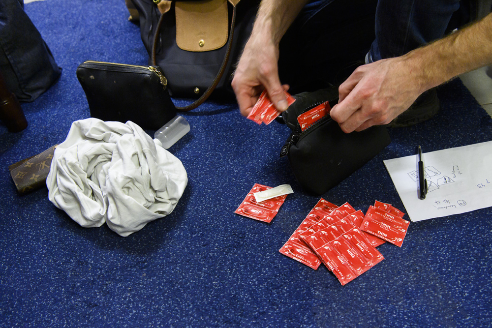 Två händer plockar upp kondomer ur en liten handväska och lägger ut i en stor samling på golvet. Foto.