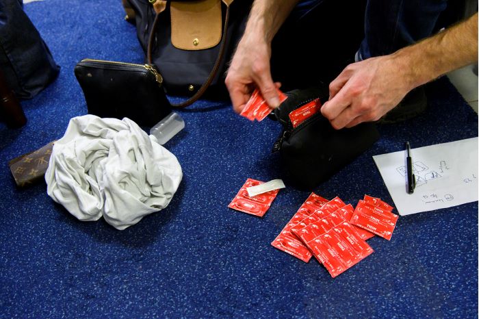 Två händer plockar upp kondomer ur en liten handväska och lägger ut i en stor samling på golvet. Foto.