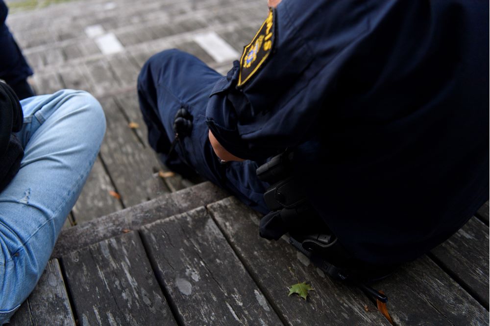 En polis i uniform sitter på en trätrappa bredvid en ungdom. Arrangerad bild.