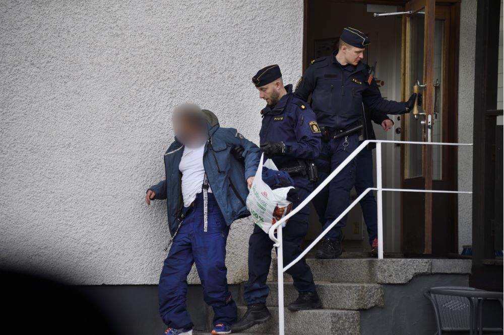 Poliserna Marcus Tengvall och Torbjörn Eliasson lotsar en man i blåa funktionskläder nedför en trappa utanför porten till ett hyreshus.
