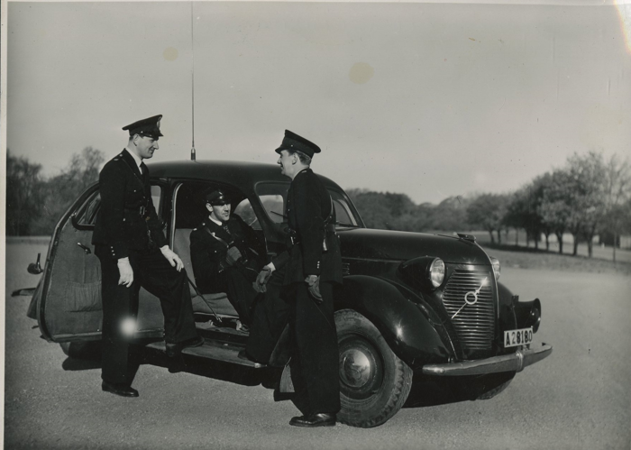 Två poliser står och pratar med varandra utanför en radiobil, i vilken ytterligare en radiopolis sitter. Foto.