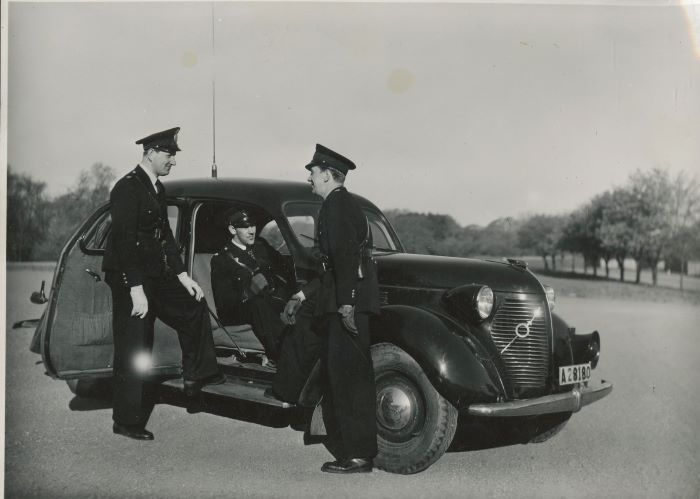 Två poliser står och pratar med varandra utanför en radiobil, i vilken ytterligare en radiopolis sitter. Foto.