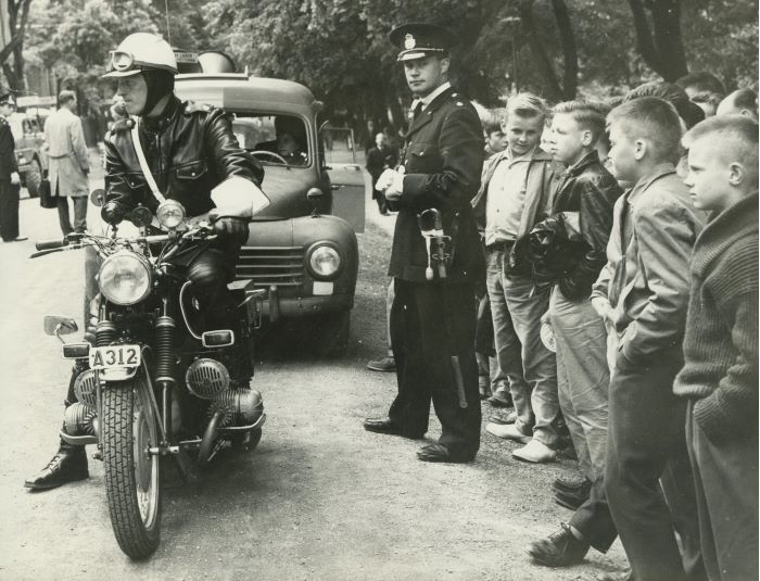 En patrullerande konstapel i polisuniform modell 1954 bevakar ordningen under en folksamling. Foto.