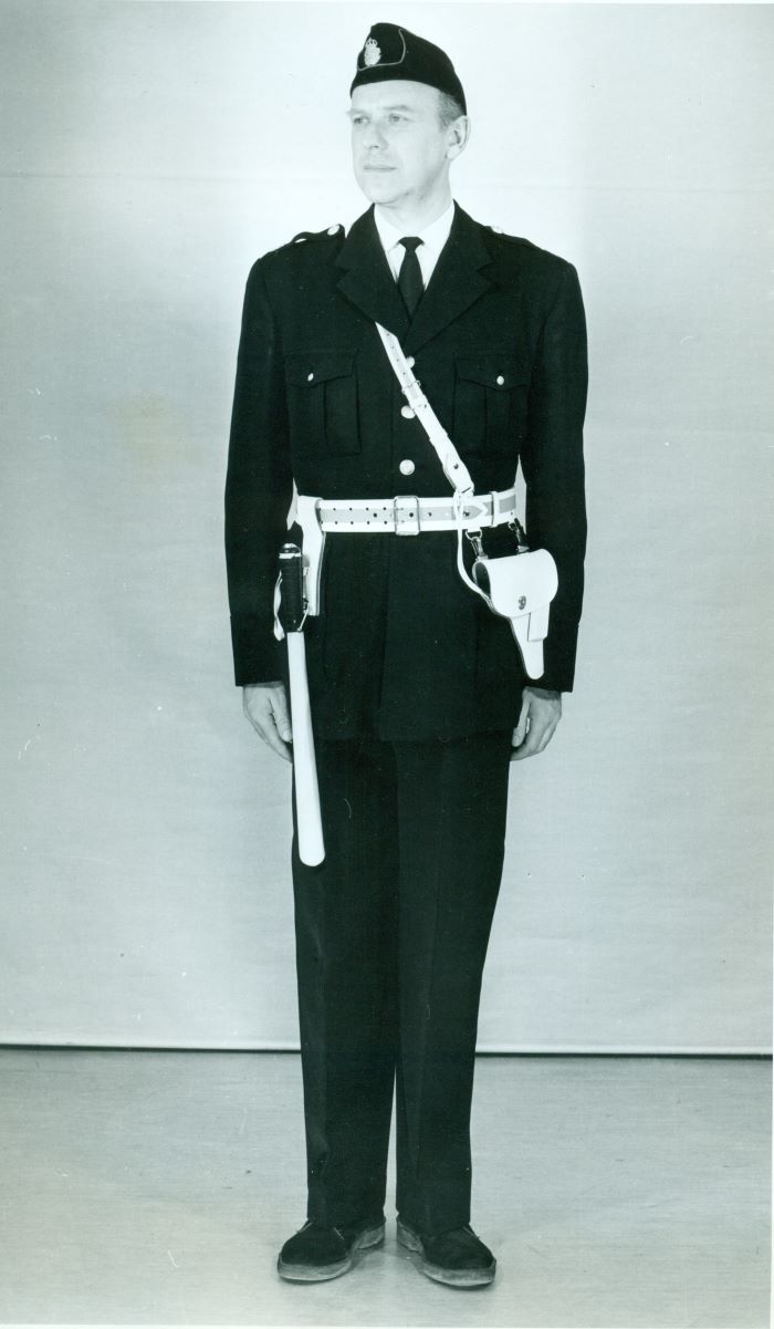En konstapel i polisuniform modell 1965, medvit batong och tjänstepistol i vitt hölster. Foto.
