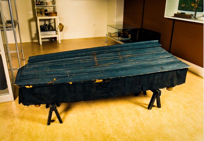 En svart gammal träkista står på ett golv i ett utställningsrum. Foto.