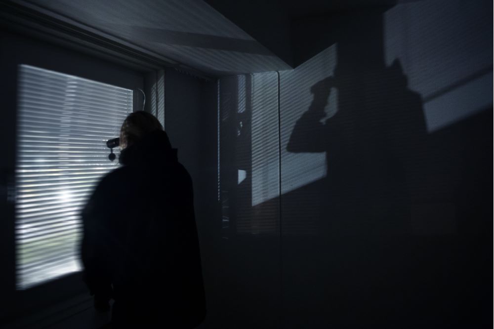 Spanaren Jennifer ses stå och titta med kikare ut genom neddragna persienner i ett helt mörkt rum. Foto.