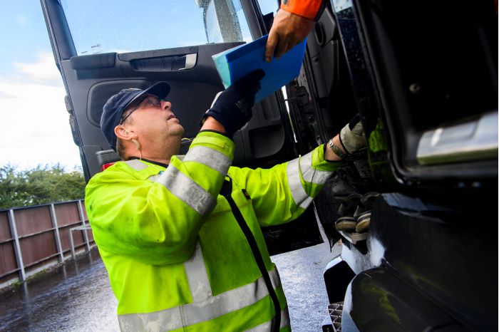 Man med reflexväst tar emot blå plastficka från en förare som sitter i en lastbil.
