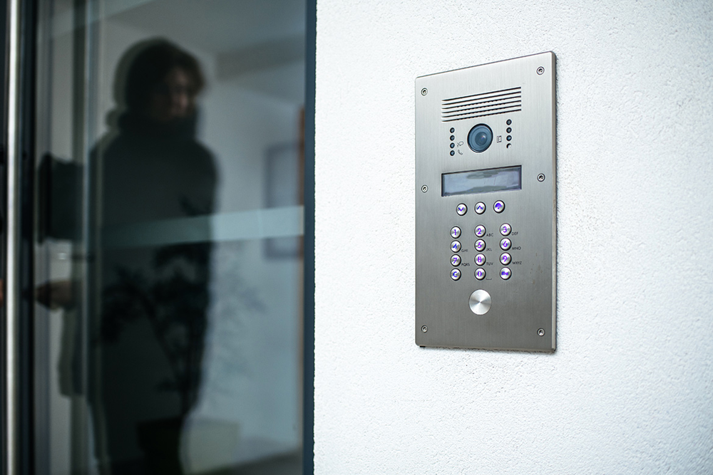 En siluett av en person speglas i fönsterglaset på en ytterdörr och bredvid syns en porttelefon med knappar. Foto.