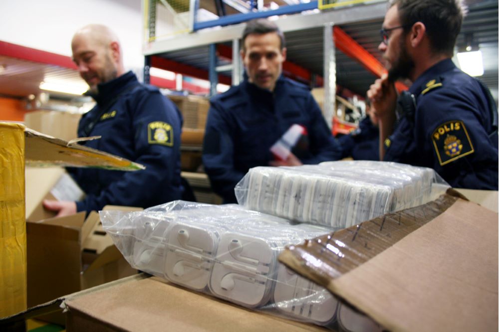En låda med ett stort antal piratkopierade hörlurar till mobiltelefon, står på ett bord i en lagerlokal. I bakgrunden syns tre poliser i uniform.