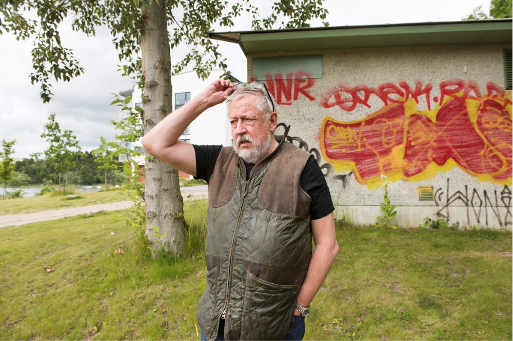 Leif GW Persson står utomhus framför en nerklottrad vägg.