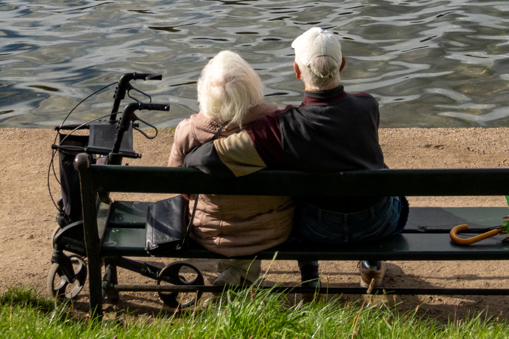 Ett äldre par sitter på en bänk och tittar ut över vattnet. En rullator står bredvid kvinnan och mannen håller om hennes axlar. Foto.