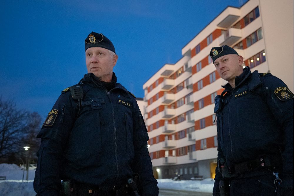 Snön ligger decimetertjock och teHenrik Herou och Adam Lofterud ur trygghetsgruppen står utomhus med ett flerfamiljshus i bakgrunden mot en mörkblå himmel. Foto.