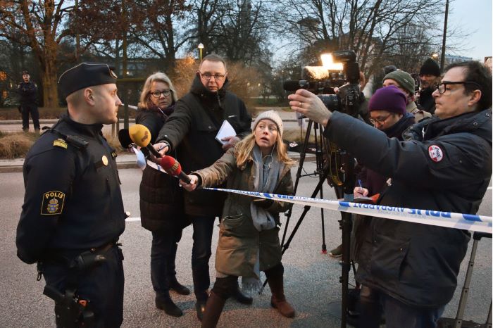 Polisen Peter Ljungsryd står bakom en avspärrning med händerna på ryggen och svarar på frågor från journalisterna som samlats på andra sidan avspärrningstejpen. Mikrofoner, lampor och filmkameror riktas från pressuppbådet mot Peter.