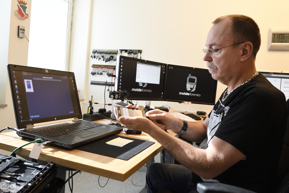 Paul Liedberg sitter vid ett skrivbord framför tre datorskärmar. I handen håller han den skjutne mannens mobiltelefon.