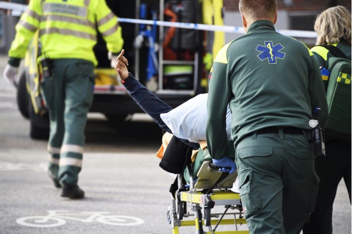 En kille och en tjej från ambulansen kör bort båren med den skadade mannen mot ambulansen. Mannen som ligger på båren sträcker ut långfingret mot fotografen.