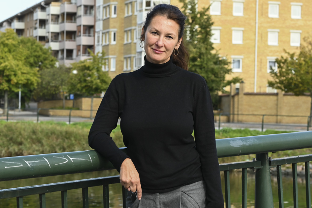 Susanne Gosenius står på en bro med lägenhetshus i bakgrunden. Foto.