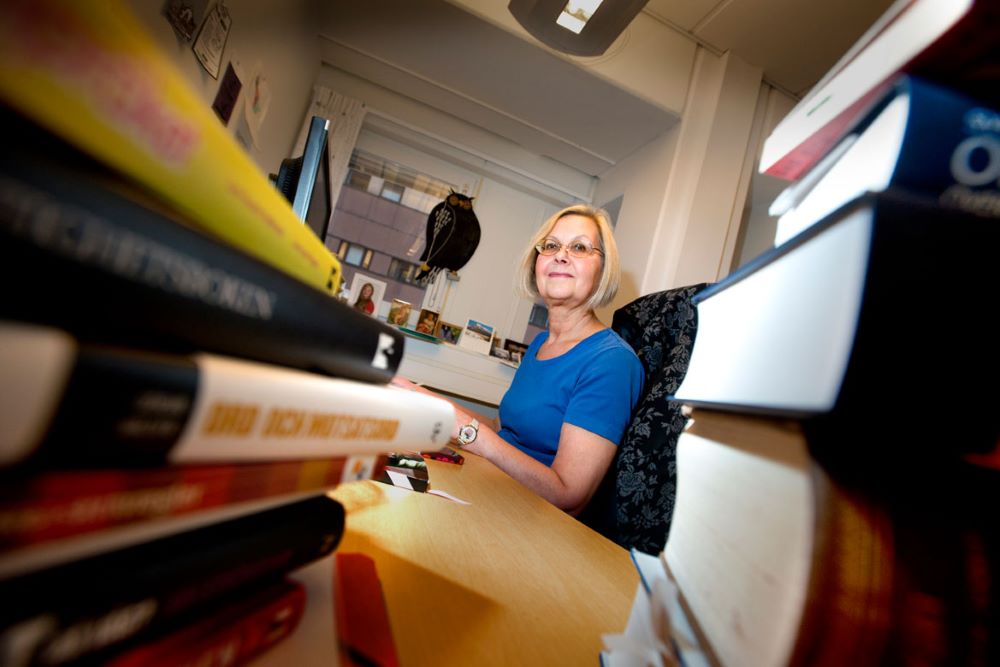 Carina Alfredsson skymtar fram vid sitt skrivbord bakom högar med böcker.