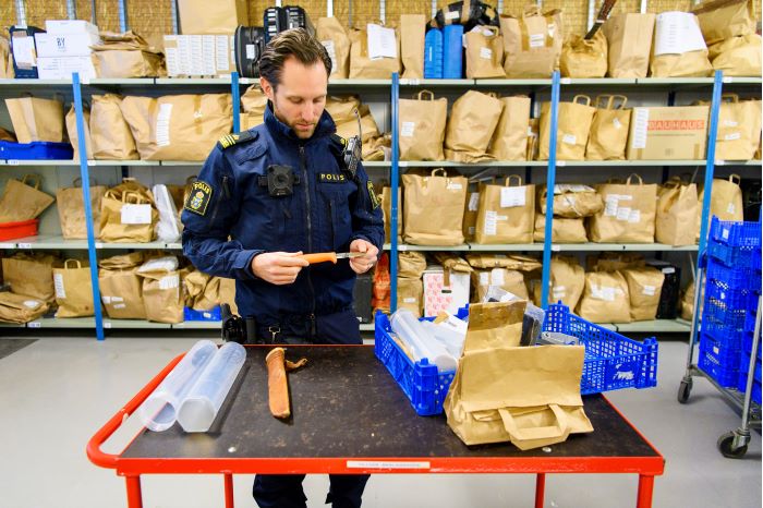 En polis håller i en hantverkarkniv som tagits i beslag. I bakgrunden finns hyllmeter med beslag i papperskassar. Foto.