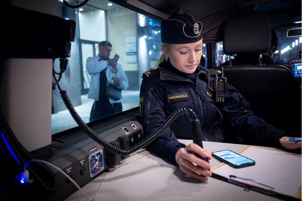 Maja Sandell sitter vid ett bord i polisbussen med en rakelenhet i handen och håller koll på gruppchatten i telefonen. Foto.