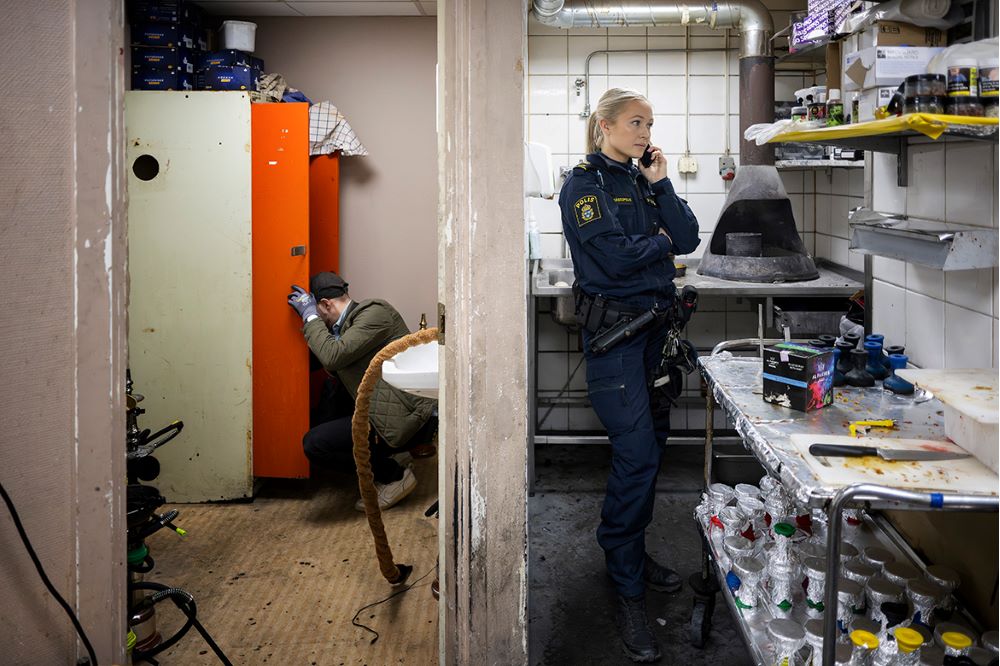 Maja Sandell står i ett källarrum i en restaurang och pratar i telefon. En civilklädd kollega letar i ett skåp i rummet bredvid. Foto.