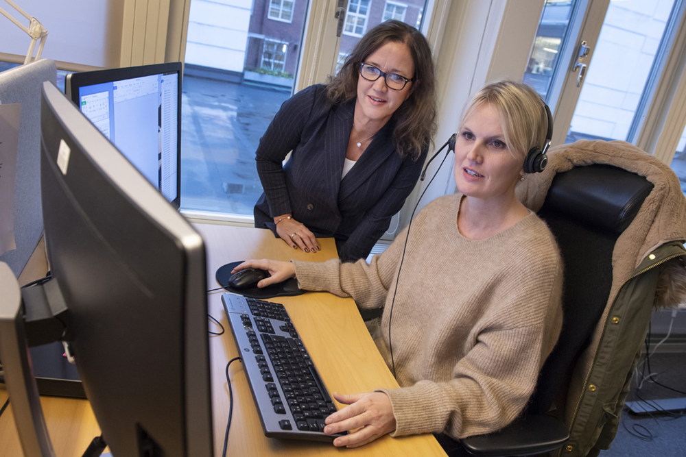 Framför två bildskärmar sitter operatör Jessica Thor med ett headset på huvudet. Förundersökningsledare Lotta Larsson står böjd över skrivbordet.
