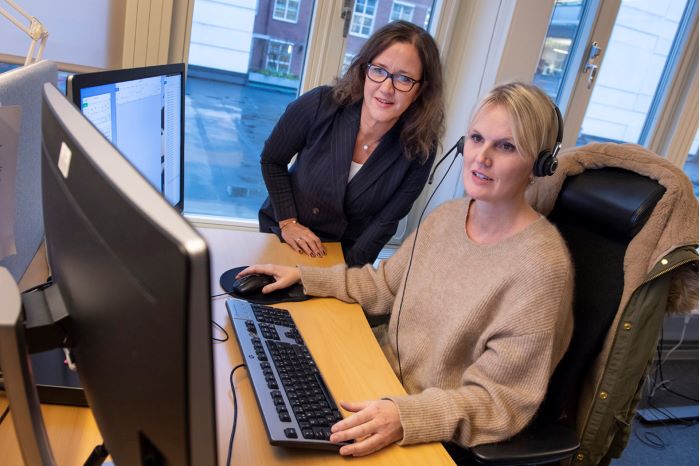 Framför två bildskärmar sitter operatör Jessica Thor med ett headset på huvudet. Förundersökningsledare Lotta Larsson står böjd över skrivbordet.