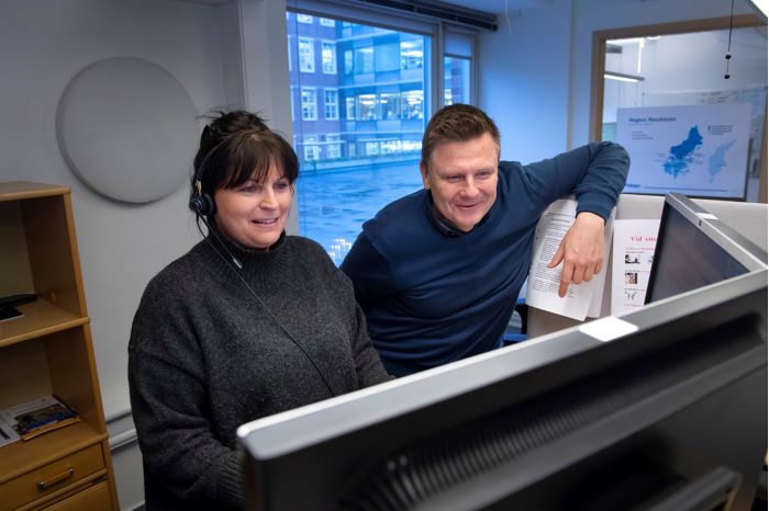 Birgitta Larsson sitter på sin arbetsplats med headset på huvudet, och tittar på en bildskärm. Bredvid står Lars Ivarsson och hänger med ena armen mot skärmskyddet vid sidan av skrivbordet.