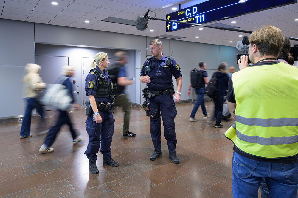 Lina och kollegan Olle framför kameran på Arlanda flygplats med resenärer i bakgrunden. Foto.