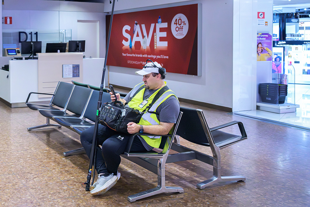 Ljudteknikern Aras sitter på en stol i rader av stolar utanför en taxfreebutik. Foto.