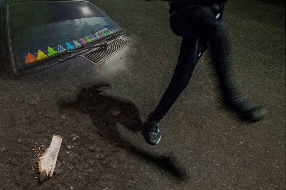 Ett fotomontage med mörk asfalt, en motorhuv med färgglada wunderbaums, en mörkklädd person som sparkar till framför sig, en sönderflisad träbit på marken.