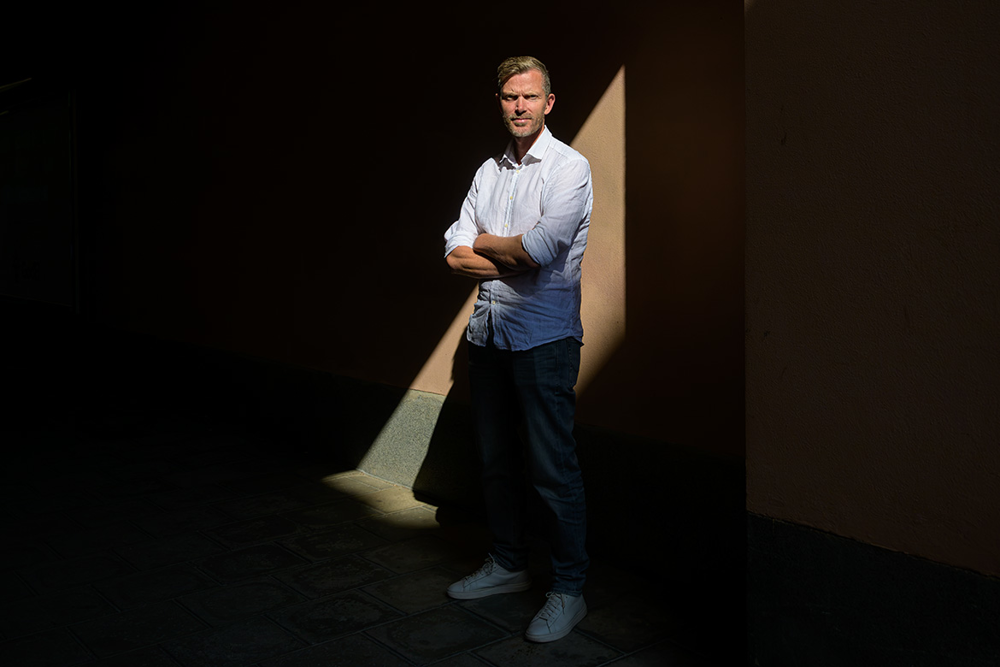 Patrik Larssson står utomhus framför en mörk vägg, där solen skiner ner och bildar mönster mot bakgrunden. Foto.