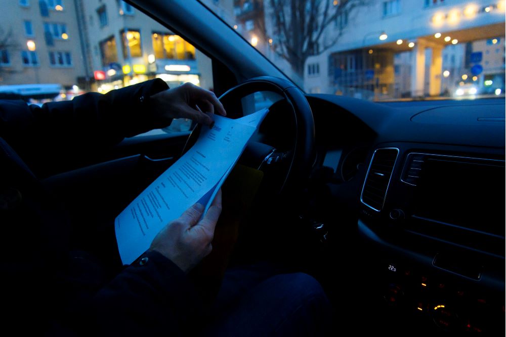 Ett par manshänder ses hålla i en bunt med papper framför ratten i en bil. Foto.