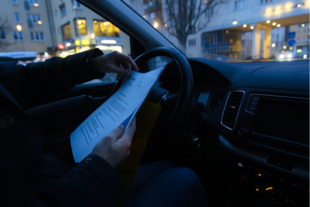 Ett par manshänder ses hålla i en bunt med papper framför ratten i en bil. Foto.