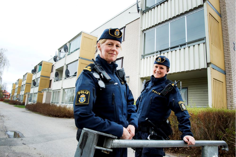 Poliserna Anna Sandberg Ställ och Marie Edlund står vid en vägbom till ett bostadsområde, med gula tegelhus i bakgrunden.