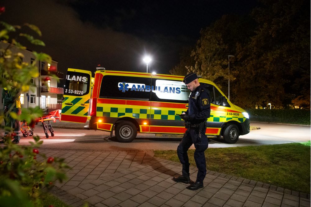 En polis ses stå på en trottoar framför en ambulans. Foto.