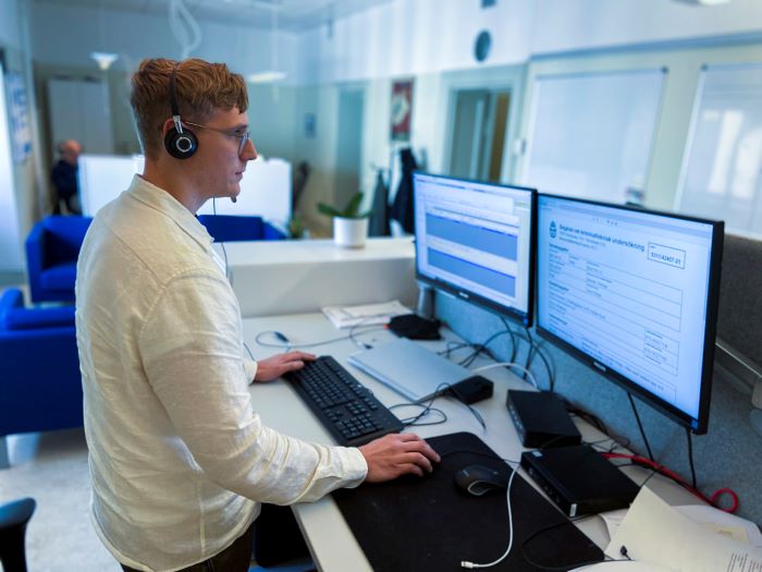 Handläggare Johan Gustafsson står vid sitt skrivbord med headset och tittar på sina datorskärmar. Foto.